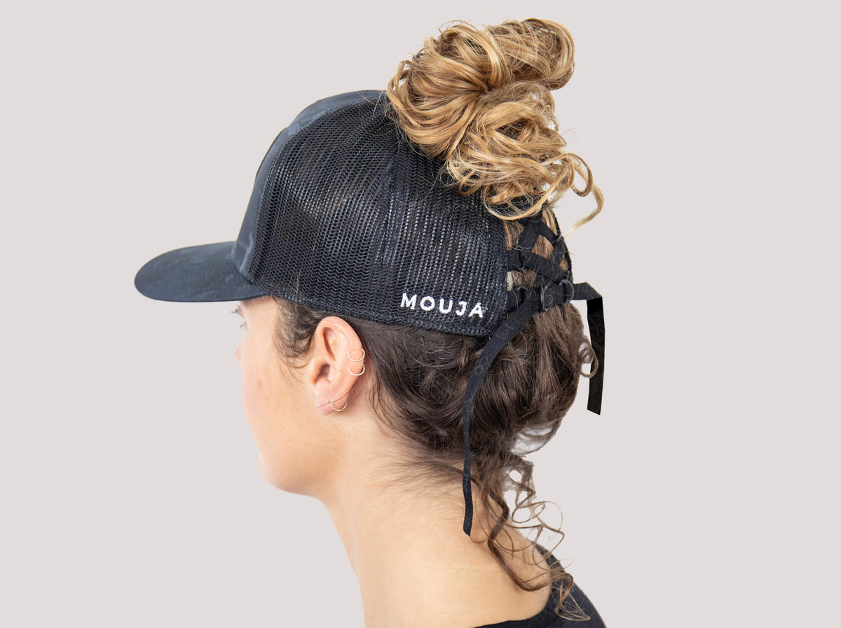 Ponytail baseball hat with black camo print. Mesh trucker style. Rear opening with cross tie. Model wearing a ponytail cap.
